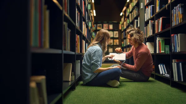 Biblioteca Universitaria: Chica inteligente sentada y niño talentoso sentado con las piernas cruzadas en el suelo, hablar, usar tableta digital y discutir el papel, estudiar y prepararse para los exámenes juntos. Estudiantes auténticos — Foto de Stock