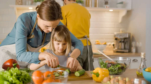 In Kitchen: Mother and Cute Little Daughter Cooking Together Healthy Dinner. Mom Teaches Little Girl Healthy Habits and how to Cut Vegetables for Salad. Cute Child Helping Her Beautiful Caring Parents
