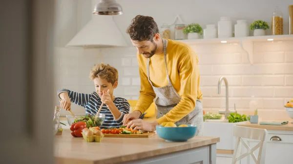 In Kitchen: Father and Cute Little Boy Cooking Together Healthy Dinner. Papá le enseña a su hijo pequeños hábitos saludables y cómo mezclar verduras en el Ensaladera. Lindo niño ayudando a sus hermosos padres cariñosos —  Fotos de Stock