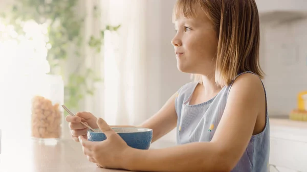 Aamiainen keittiössä: Suloisen pienen tytön muotokuva syö terveellistä granolaa muroviljaa maidon kanssa kulhosta. Söpö pikkutyttö aamiaisella. Taustassa huolehtiva äiti valmistaa ruokaa — kuvapankkivalokuva
