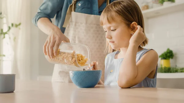 Pequeno-almoço na Cozinha: Jovem Mãe Bonita Despeja Cereais em Tigela, Filha Adorável Preparando-se para Comer com Prazer. Mãe carinhosa prepara café da manhã de cereais para sua linda menina — Fotografia de Stock