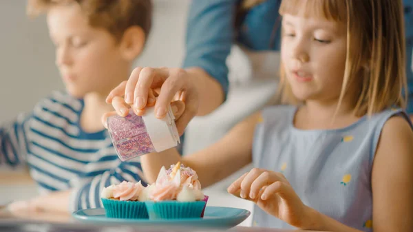 In the Kitchen: Portrait of the Cute Little Daughter Sprinkling Funfetti on Creamy Cupcakes Frosting. Family Cooking Muffins Together. Adorable Children Helping their Caring Parents — Stock Photo, Image