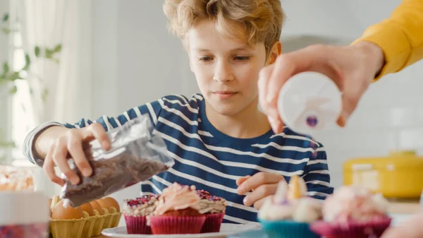 In the Kitchen: Portrait of the Smart Little Boy Sprinkling Funfetti on Creamy Cupcakes Frosting. Família Cozinhar Muffins Juntos. Adoráveis crianças ajudando seus pais carinhosos — Fotografia de Stock