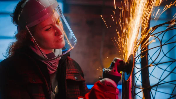 Joven artista femenina contemporánea está moliendo su escultura de tubo de metal con una herramienta eléctrica portátil en un taller de estudio. Empoderar a la mujer hace obras de arte abstractas brutales modernas de metal . — Foto de Stock