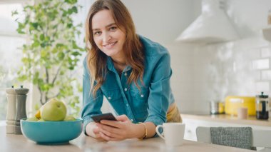 Güzel Kadın, Mutfakta Akıllı Telefon Kullanıyor 'muş. Kameraya Güzel Genç Kadın Poz Veriyor ve Nazikçe Gülümsüyor. Sağlıklı Yaratıcı Yaşam Tarzı olan Çekici Kız Sabahları Evde Rahatlıyor.