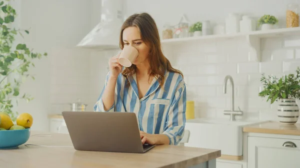 青いパジャマを着ている間、ラップトップコンピュータを使用し、コーヒーや紅茶の朝のカップを飲む若い美しい女性。現代的なサニーキッチンルームに座っているブルネットの女性。フリーランス｜自宅で働く. — ストック写真