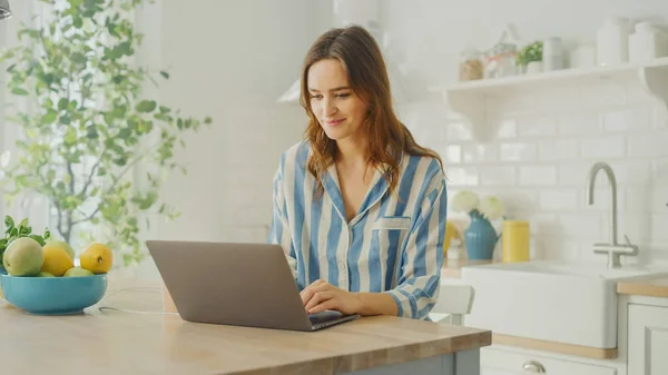 Mulher bonita nova usando computador portátil enquanto vestindo pijama azul. Morena Feminino Sentado em uma sala de cozinha moderna. Trabalhando em casa durante a quarentena . — Fotografia de Stock