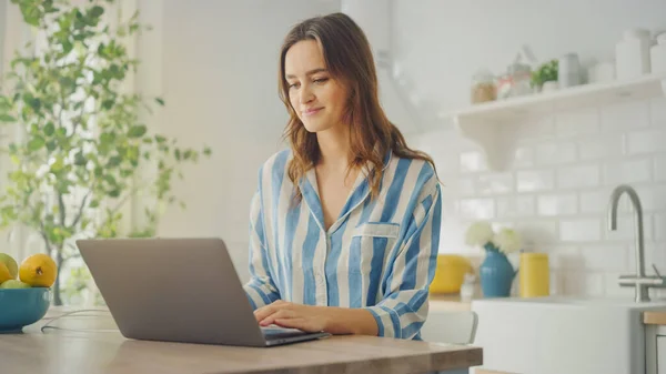 青いパジャマを着ている間、ラップトップコンピュータを使用して若い美しい女性。現代的なキッチンルームに座っているブルネットの女性。検疫中に自宅から働く. — ストック写真
