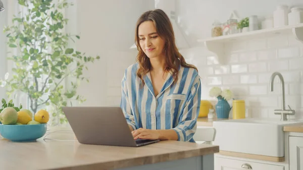 Mulher bonita nova usando computador portátil enquanto vestindo pijama azul. Morena Feminino Sentado em uma moderna sala de cozinha ensolarada. Freelancer Trabalhando em casa durante a quarentena . — Fotografia de Stock