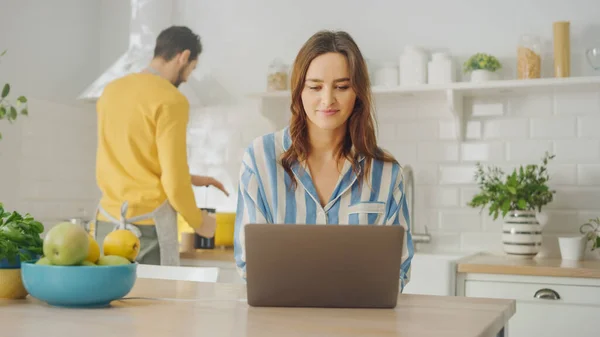 Молода красива жінка використовує ноутбук комп'ютер під час носіння блакитної піжами. Брюнетка жінка сидить у сучасній сонячній кухні. Фрілансер Робота з дому . — стокове фото