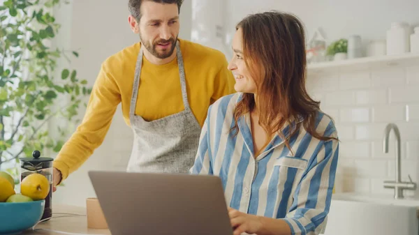 Hermosa pareja joven usando ordenador portátil en la cocina en casa. Mujer con pijama. El hombre que usa un delantal está ayudando a su novia con el trabajo en línea. Mujer sentada y navegando por Internet . —  Fotos de Stock