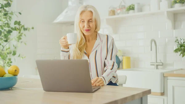 Mulher Sênior bonita usando computador portátil e beber uma xícara de café ou chá em uma cozinha ensolarada. A avó está conectada com as crianças via internet e mídias sociais. Pensionista trabalhando a partir de casa . — Fotografia de Stock