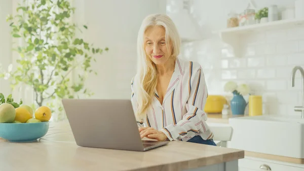 Mulher Sênior bonita usando computador portátil em uma cozinha ensolarada. Ela sorri. A avó está conectada com as crianças via internet e mídias sociais. Pensionista trabalhando a partir de casa . — Fotografia de Stock