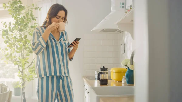 아름답게 생긴 여성 이 프리큐어를 한 잔의 커피를 마시면서 부엌에서 스마트 폰을 사용하고 있다. 아침에 집에서 건강에 좋은 생활 방식을 유지 한 파이오니아 소녀. — 스톡 사진