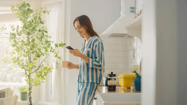 아름답게 생긴 여성 이 프리큐어를 한 잔의 커피를 마시면서 부엌에서 스마트 폰을 사용하고 있다. 아침에 집에서 건강에 좋은 생활 방식을 유지 한 파이오니아 소녀. — 스톡 사진