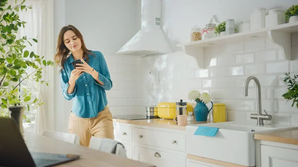 Приваблива молода жінка в джинсах Шерт і Браун Штанці використовує свій смартфон на кухні. Вона танцює в сучасній кухні з здоровим стилем життя. Щаслива прекрасна дівчинка відпочиває вдома. — стокове фото