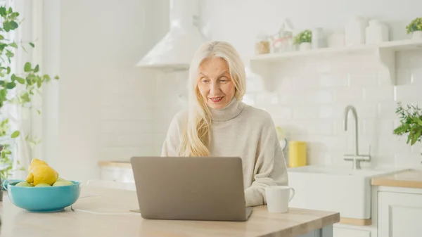 Hiteles Senior Nő Laptop Számítógépet használ egy fényes konyhaszobában otthon. Gyönyörű öreglány nyugdíjas ősz hajjal Böngészés Internet a számítógépen. Boldog idős ember, tele egészséggel. — Stock Fotó