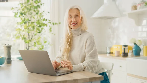 Gyönyörű idős nő Laptop számítógépet használ egy napfényes konyhában. A kamerába és a mosolyba néz. Nagymama kapcsolódik a gyermekek az interneten és a közösségi média. Nyugdíjas Otthonról dolgozik. — Stock Fotó