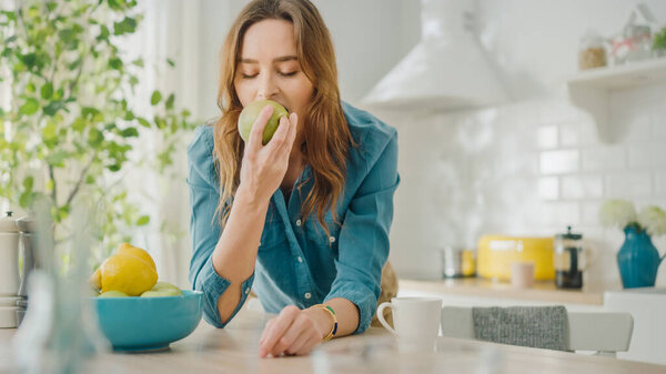 Authentic Beauty Portrait of a Beautiful Brunette Female Eating a Big Green Apple in a Bright Room at Home. Pretty Young Woman Poses for the Camera and Gently Smiles. Healthy Natural Vegetarian.