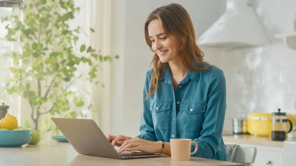 "Молода приваблива жінка" використовує Laptop Computer and Drinking Morning Cup of Coffee, одягаючи джинсову сорочку. "Brunette Female Sitting in a Modern Cozy Kitchen"). Фрілансер, що працює вдома. — стокове фото