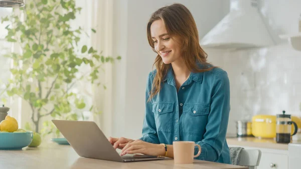 Jovem Mulher Atraente Usando Computador Laptop e Bebendo Copa Manhã de Café Enquanto Vestindo uma Camisa Jeans. Morena Feminino Sentado em uma cozinha moderna acolhedora. Freelancer Trabalhando em casa . — Fotografia de Stock