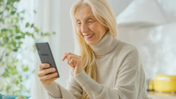 Auténtica mujer mayor usando Smartphone en la sala de cocina en casa. Hermosa anciana pensionista con pelo gris utiliza una aplicación en un teléfono celular. Persona de edad avanzada en manos de la tecnología . — Foto de Stock