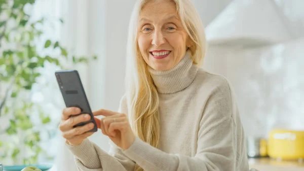 自宅のキッチンルームでスマートフォンを使用して本格的なシニア女性。白髪の美しい古い女性年金受給者は、携帯電話上のアプリケーションを使用しています。高齢者はカメラと笑顔を見て. — ストック写真