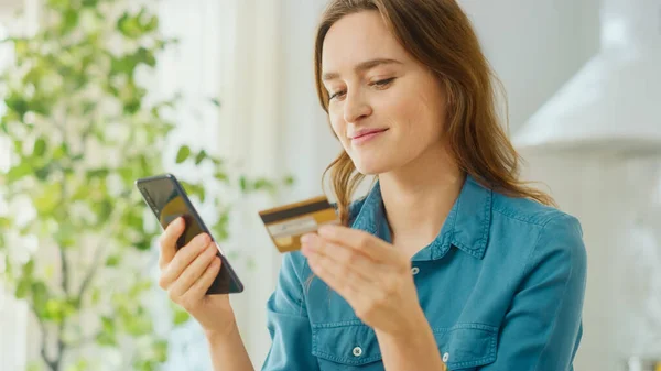 Vacker kvinna Använda smartphone i ett ljust soligt kök. Hon betalar en faktura för onlineköp med sitt kreditkort. Glad attraktiv flicka med hjälp av e-handel program på hennes mobil. — Stockfoto