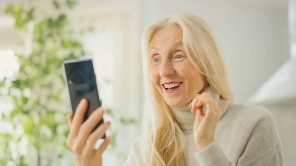 Authentic Senior Woman Respondiendo una videollamada usando su teléfono inteligente en la sala de cocina en casa. Hermosa anciana pensionista con el pelo gris está feliz de ver a su familia en el chat de vídeo . — Foto de Stock