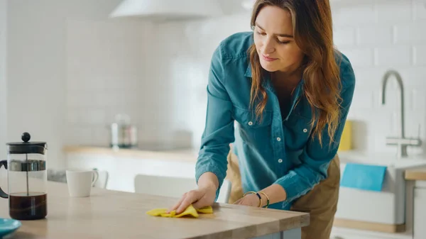 Caring Housewife Wiping Spilled Coffee or Crumbs from a Wooden Kitchen Table. Beautiful Young Female Using a Household Cleaner Wipe for Cleaning. Brunette is Wearing a Jeans Shirt and Beige Pants.