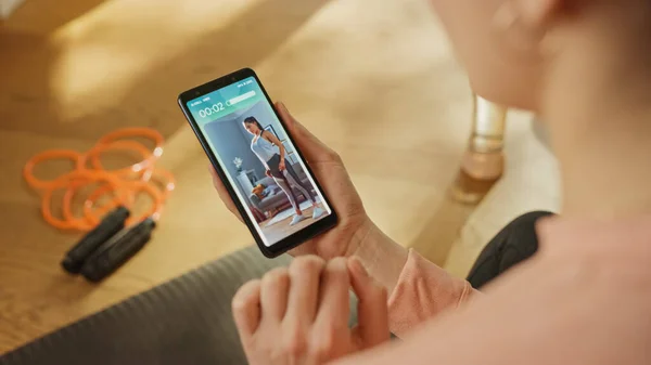 Over Shoulder Shot: Fit Woman Holds Smartphone, utilizza l'app di monitoraggio del fitness, guardando video premium che spiega l'esercizio. Software di applicazione di formazione finto — Foto Stock
