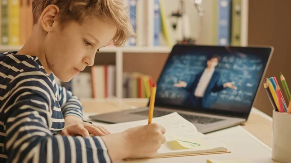 Смарт-маленький хлопчик використовує ноутбук для відеодзвінків зі своїм вчителем. Екран показує онлайн-лекцію з вчителем, що пояснює тему з класу. Замкнене дистанційне навчання, домашнє навчання — стокове фото