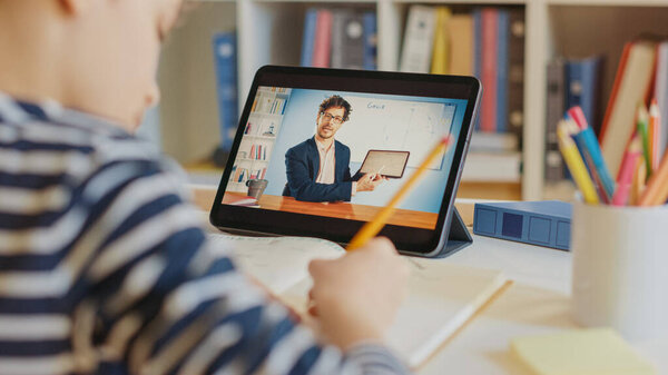 Little Boy Uses Digital Tablet for Video Call with His Teacher. Screen Shows Online Lecture with Teacher Explaining Subject from Classroom. E-Education Distance Learning, Homeschooling. Over Shoulder