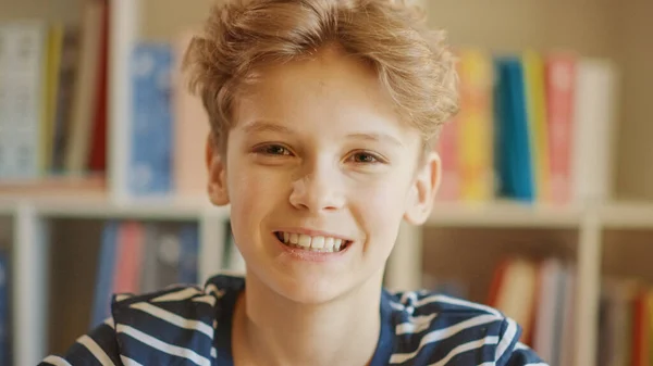 Närbild Porträtt av en glad smart ung pojke, leende och tittar på kameran. Bakgrund suddig bokhylla. — Stockfoto