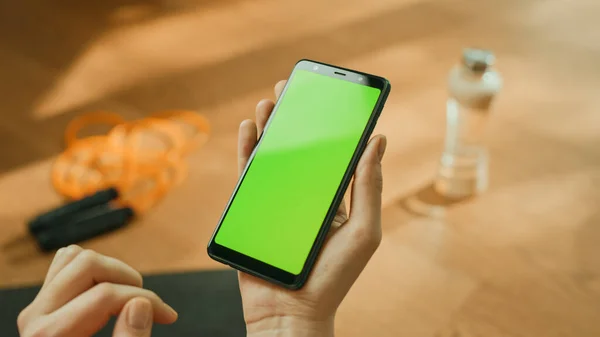 Fitness koncepció: nő kezében Chroma Key Green Screen Smartphone. A háttérben Fitness matrac, ugráló kötél és felszerelés. Közelkép a nézőpontról POV — Stock Fotó