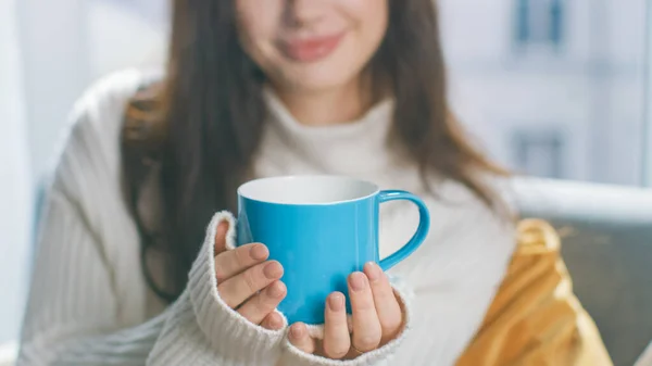 Syuting Young Brunette Holding Blue Cup dengan Hot Beverage. Sweet Girl Wearing White Knitted Sweater, Spends Relaxing Time by Herself In Cozy Living Room — Stok Foto