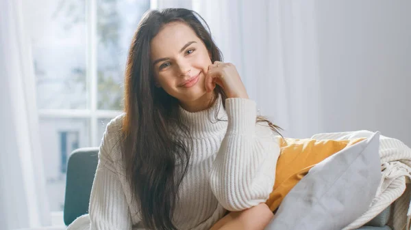 Shot of Beautiful Young Brunette Smiling Charmingly while Looking at Camera. Shy Girl Wearing White Knitted Sweater Laughing in Cozy Room