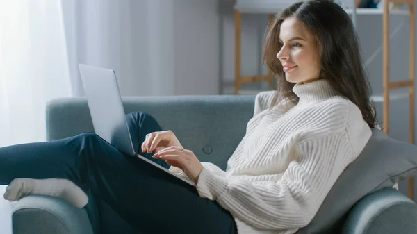 Mulher bonita trabalha no computador portátil enquanto sentado na cadeira. Sensual menina vestindo camisola funciona em estudos de caderno, Surfs Internet, usa as mídias sociais enquanto relaxa no aconchegante apartamento — Fotografia de Stock