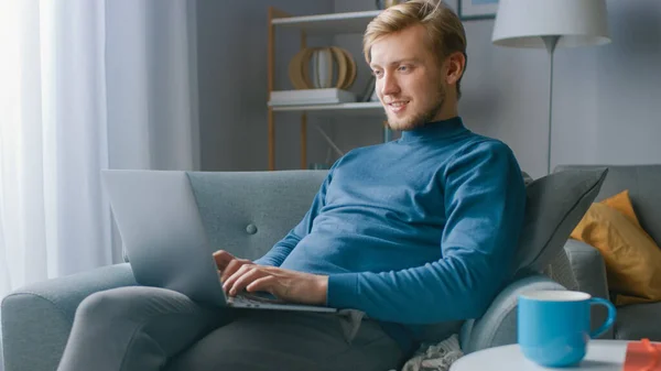 Πορτρέτο του Όμορφου Ξανθού Νέου που εργάζεται σε έναν φορητό υπολογιστή, ενώ κάθεται σε μια καρέκλα στο άνετο σαλόνι του. Creative Freelancer Relaxes at Home, Surfs Internet, Χρησιμοποιεί τα Social Media και Relaxes — Φωτογραφία Αρχείου