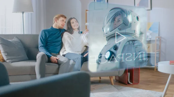 Krásná mladá dvojice sedí na gauči doma a vybírá film, který se bude dívat na futuristickou holografickou obrazovku. — Stock fotografie