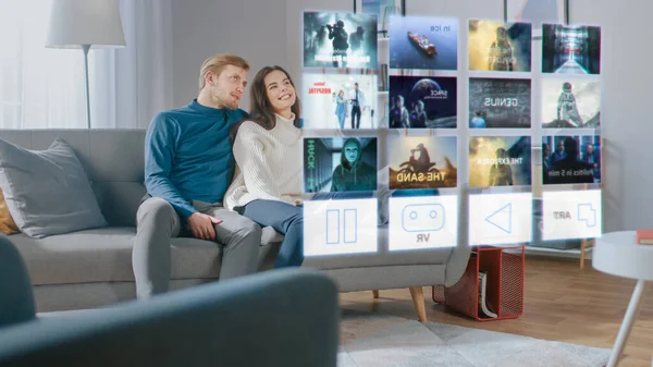 Hermosa pareja joven se sienta en un sofá en casa y elegir una película para ver en una pantalla holográfica futurista . — Foto de Stock