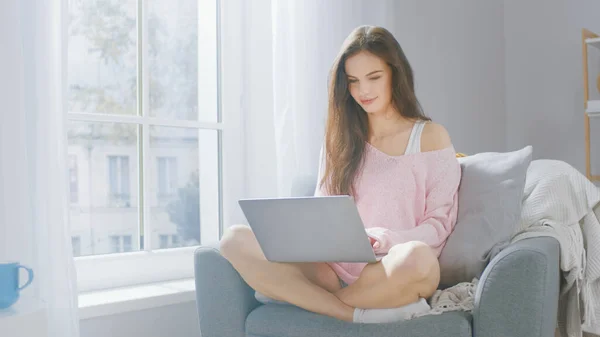 Retrato de Jovem Morena Funciona em Computador portátil em casa em uma sala de estar acolhedora. Menina lindo vestindo camisola rosa, gasta tempo relaxante . — Fotografia de Stock