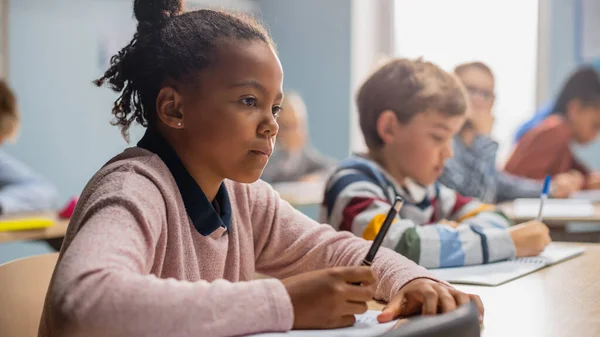 Im Klassenzimmer der Grundschule schreibt ein brillantes schwarzes Mädchen in einem Aufgabenheft, macht Tests und schreibt Prüfungen. Juniorklasse mit vielfältiger Gruppe von Kindern, die fleißig arbeiten und Neues lernen — Stockfoto