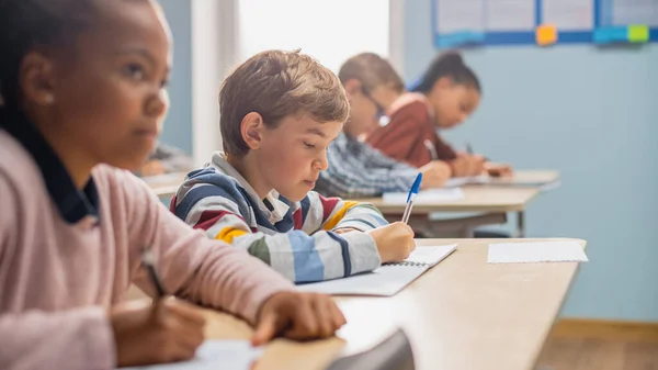 In Elementary School Classroom Brilliant Black Girl Writes in Exercise Notebook, Taking Test and Writing Exam. Junior Classroom with Diverse Group of Children Working Diligently and Learning New Stuff — Stock Photo, Image