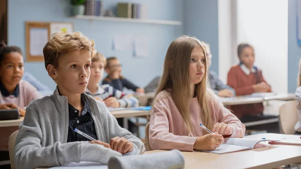 Sala de aula elementar de várias crianças ouvindo atentamente a sua lição de dar professor. Jovens brilhantes na escola escrevendo em cadernos de exercícios, fazendo testes, exames. Criança com braço quebrado . — Fotografia de Stock