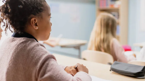 In Elementary School Class: Over the Shoulder View of a Brilliant Black Girl Listening to a Teacher. Младший класс с разношерстной группой ярких детей, работающих и создающих новые вещи — стоковое фото