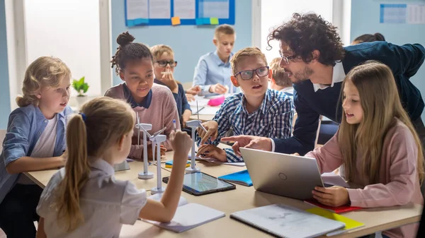 Aula de la escuela primaria: Profesor entusiasta sosteniendo la tableta La computadora explica a un niño pequeño brillante cómo funcionan las turbinas de viento. Niños aprendiendo sobre las formas ecológicas de energía renovable — Foto de Stock