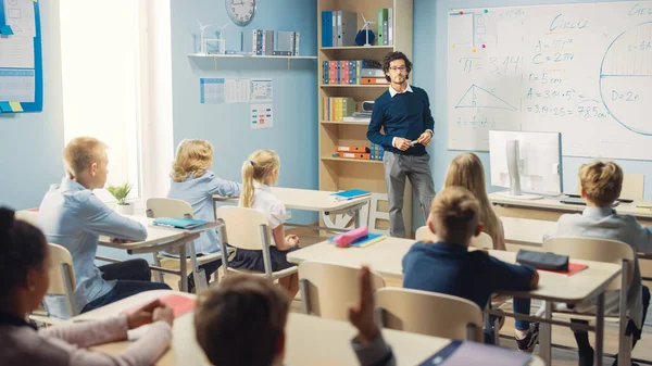 Tehetséges tanár elmagyarázza a leckét egy osztályteremnek, ami tele van ragyogó, változatos gyerekekkel. Az általános iskolában az intelligens többnemzetiségű tanulás geometriája és fizikája, fiú felemelő kéz egy választ — Stock Fotó