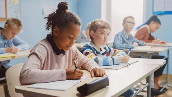 Dalam Elementary School Classroom Brilliant Black Girl Writes in Exercise Notebook, Taking Test and Writing Exam (dalam bahasa Inggris). Kelas Junior dengan Kelompok Diverse Anak-anak Bekerja Diligen dan Belajar Barang Baru — Stok Foto