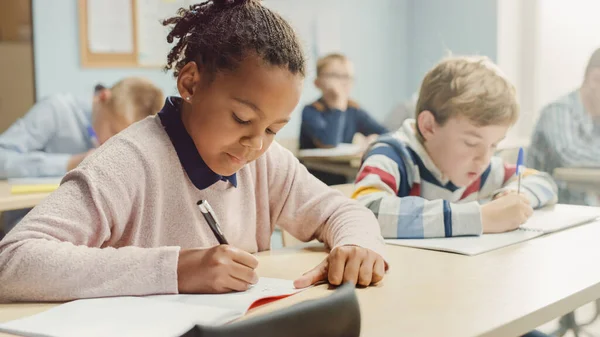 Dalam Elementary School Classroom Brilliant Black Girl Writes in Exercise Notebook, Taking Test and Writing Exam (dalam bahasa Inggris). Kelas Junior dengan Kelompok Diverse Anak-anak Bekerja Diligen dan Belajar Barang Baru — Stok Foto